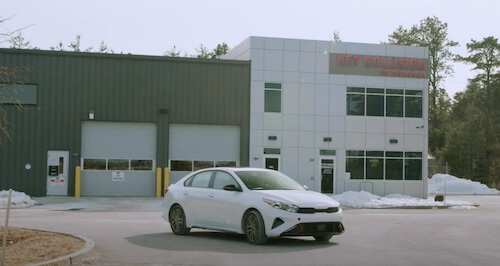 A white sedan car is parked in front of a building with the sign 'KEY COLLISION OF CONCORD'. The building has a modern look with a mix of gray siding and white panels, and a few snow piles on the side indicate a winter or early spring season. This setting suggests the car may be ready for pick-up after servicing at the collision center.