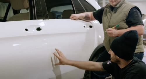 Two auto workers are focused on a task with a white car. One, with tattooed arms, is standing and holding a pen, possibly assessing or planning the work to be done. The other, wearing a beanie, is crouched and appears to be feeling the surface of the car door, possibly checking the work done or preparing for the next step.