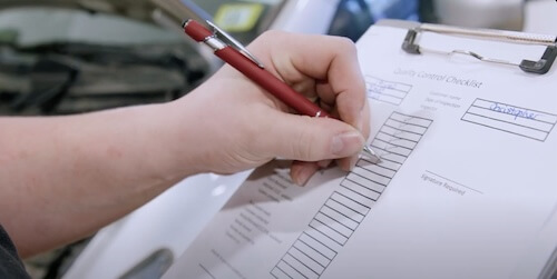 A person is filling out a quality control checklist with a red pen. The form is clipped to a board, with the name 'Christopher' filled in a field at the top. The focus is on the hand and the checklist, highlighting a step in the quality assurance process.