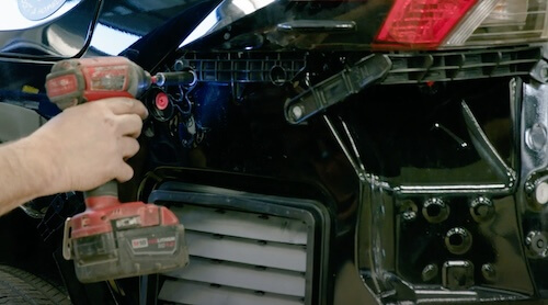 A person is using a red cordless impact driver to work on the inner mechanics of a car door, which appears to be partially disassembled. The focus is on the tool and the hand of the technician, showing a detail of the reassembly process in vehicle repair.