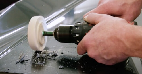A close-up of hands using a power drill with a polishing attachment on the surface of a car, creating a swirl of dust and debris. This suggests a step in the vehicle repair process, possibly buffing or sanding a section of the car body.